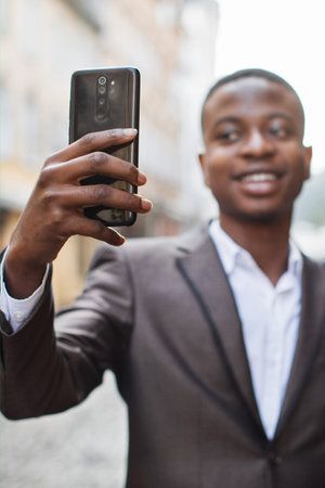 A young man in a suit jacket and white shirt smiles while holding up a smartphone to take a selfie on a city streetの写真素材