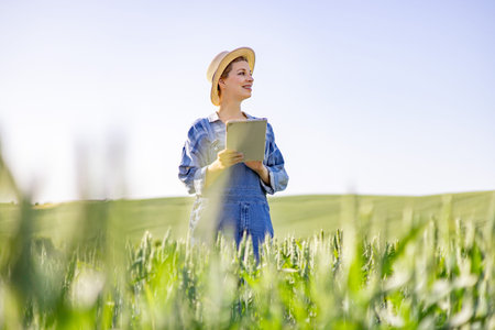 A young woman wearing overalls and a straw hat stands in a green wheat field, holding a tablet computer and looking into the distanceの写真素材
