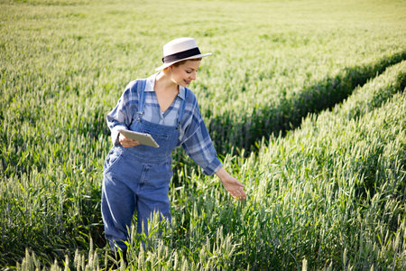 A woman in overalls and a hat uses a tablet to examine crops in a vast green field under sunlight