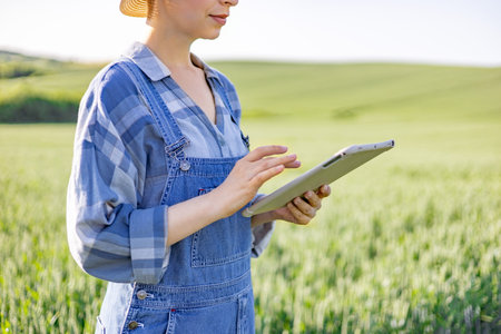 A woman in overalls and a straw hat uses a tablet computer while standing in a green agricultural fieldの写真素材