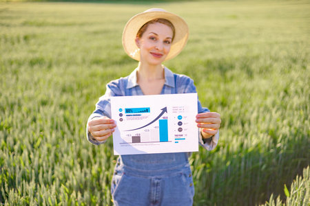 A woman in overalls and a straw hat stands in a green field holding a paper with a bar graph and an upward trending arrow indicating growthの写真素材