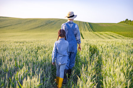 A farmer wearing overalls and a hat walks with a young child through a vast field of ripening wheat under a clear skyの写真素材