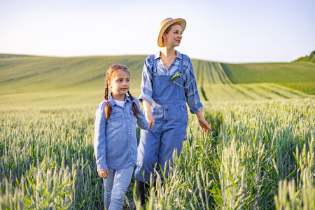 A mother and her young daughter walk hand-in-hand through a lush green wheat field under a clear sky, embodying a sense of rural life and family connectionの写真素材