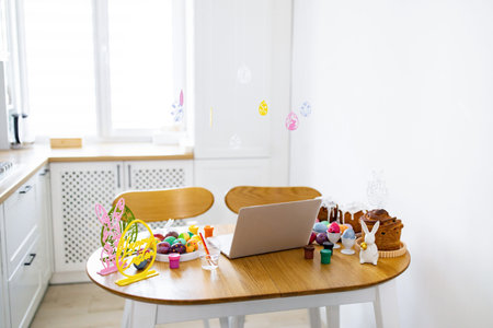 A laptop sits on a wooden table surrounded by colorful Easter eggs, paints, brushes, and traditional Easter cakes, suggesting an online celebration or craft sessionの写真素材