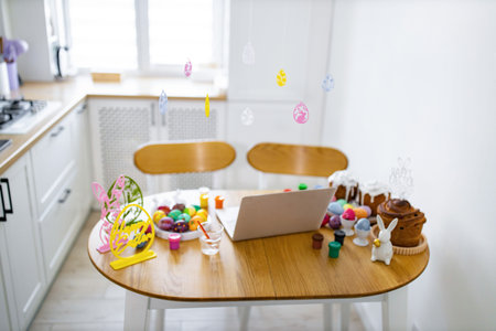 A kitchen table is set up for Easter egg decorating with painted eggs, cakes, and a laptop for virtual celebrationの写真素材