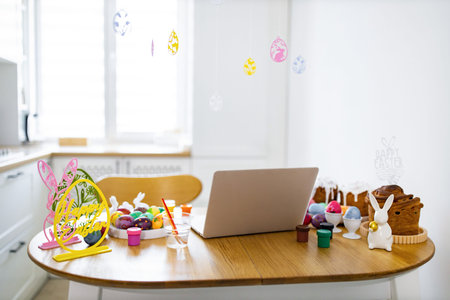 A table is set up for Easter egg decorating with paints, brushes, colorful eggs, and traditional cakes, alongside a laptop for a modern twist on holiday preparationsの写真素材
