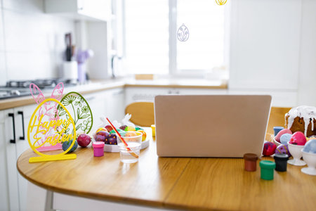 A table is set for Easter egg decorating with painted eggs, paints, brushes, and a laptop, suggesting a virtual celebration or tutorialの写真素材