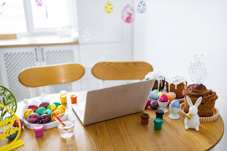 A laptop sits on a wooden table amidst colorful Easter eggs, paints, and traditional cakes, suggesting a virtual celebration or craft sessionの写真素材