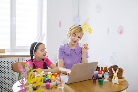 A mother and daughter wearing bunny ears are painting Easter eggs and looking at a laptop, celebrating the holiday together at homeの写真素材