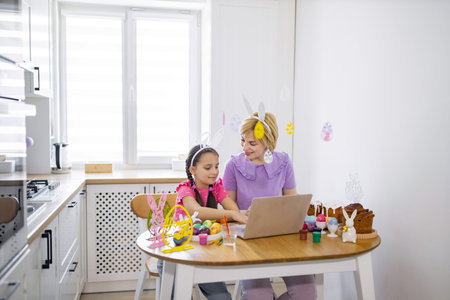A mother and daughter wearing bunny ears are engaged in an Easter activity at a kitchen table, using a laptop for inspiration or connectionの写真素材