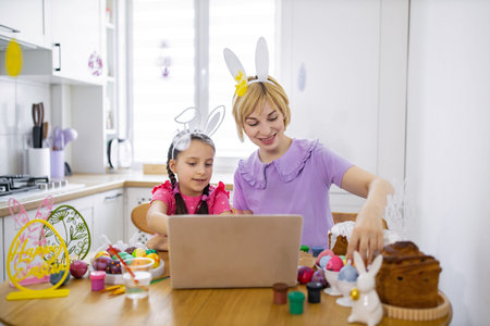 A mother and daughter wearing bunny ears are decorating Easter eggs and cakes while looking at a laptop in a bright kitchenの写真素材