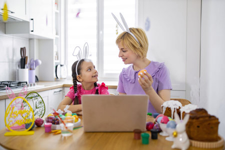 A mother and daughter wearing bunny ears are decorating Easter eggs together in a bright kitchen, with a laptop open on the tableの写真素材