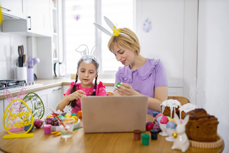 A mother and daughter wearing bunny ears are decorating Easter eggs together while watching a tutorial on a laptop in their kitchenの写真素材