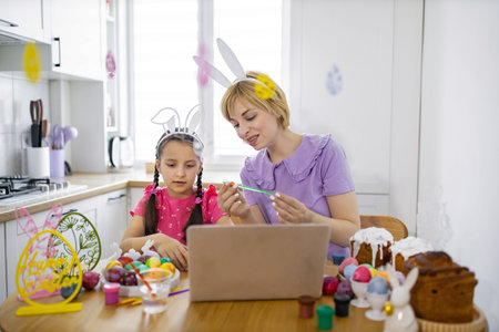 A mother and daughter wearing bunny ears are painting Easter eggs together at a kitchen table while looking at a laptop screenの写真素材