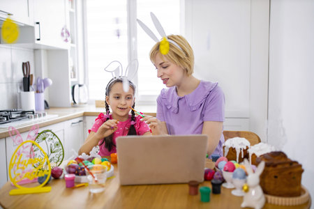 A mother and daughter wearing bunny ears participate in an online Easter egg decorating class, using a laptop for instruction and enjoying festive treatsの写真素材