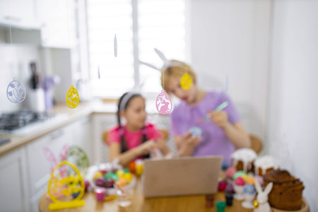 A mother and daughter are engaged in decorating Easter eggs together at a kitchen table, surrounded by festive decorations and baked goodsの写真素材