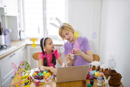 A mother and daughter wearing bunny ears engage in an Easter craft activity at home, using a laptop for inspiration or guidanceの写真素材