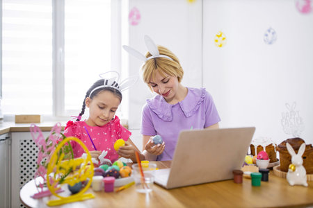 A mother and daughter wearing bunny ears paint Easter eggs together at a table while looking at a laptop screen, celebrating the holidayの写真素材