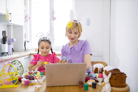 A mother and daughter wearing bunny ears are painting Easter eggs together in a bright kitchen while looking at a laptop screenの写真素材