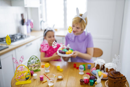 A mother and daughter enjoy decorating Easter eggs together in a bright kitchen, surrounded by festive decorations and cakesの写真素材
