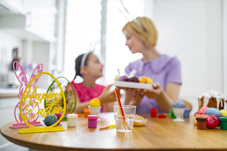 A mother and daughter wearing bunny ears are happily decorating Easter eggs together at a wooden table filled with craft supplies and painted eggsの写真素材