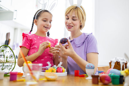 A mother and daughter wearing bunny ears are happily decorating Easter eggs together at a table...
