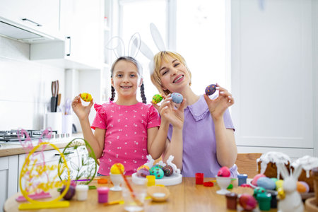 A mother and daughter wearing bunny ears happily display their decorated Easter eggs in a bright...