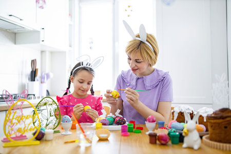 A mother and her daughter wearing bunny ears are happily painting colorful Easter eggs together at a...