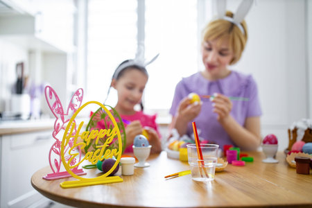 A mother and daughter happily paint colorful Easter eggs together at a kitchen table, surrounded by...