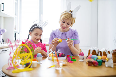 A mother and her daughter wearing bunny ears are happily painting colorful Easter eggs together at a wooden table, surrounded by festive decorations and cakesの写真素材