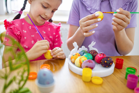 A young girl and an adult are happily painting colorful Easter eggs together at a wooden table, surrounded by paints and decorationsの写真素材