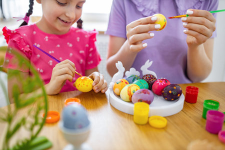 A young girl and an adult are happily painting colorful Easter eggs together at a wooden table with various paints and brushesの写真素材