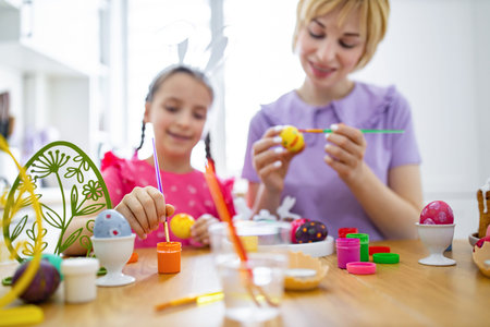 A mother and daughter happily paint colorful Easter eggs together at a wooden table, creating festive holiday decorationsの写真素材