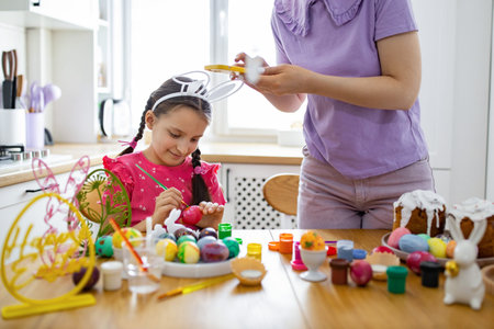 A young girl with bunny ears paints Easter eggs at a table with her mother, surrounded by festive...