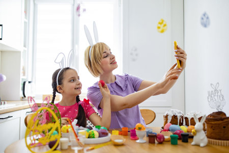 A mother and daughter wearing bunny ears take a selfie with painted Easter eggs and cakes on the...