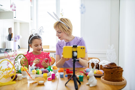 A mother and daughter wearing bunny ears paint Easter eggs together at a kitchen table while...