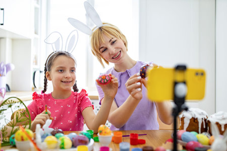 A mother and daughter wearing bunny ears are happily decorating Easter eggs and recording the...