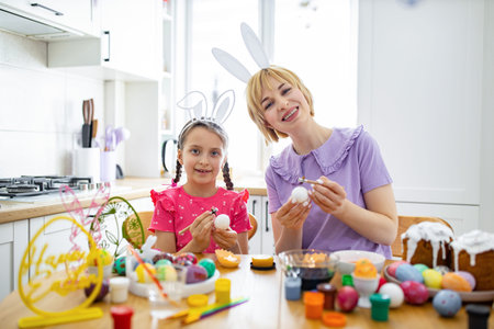 A mother and daughter wearing bunny ears happily decorate Easter eggs together at a table filled...