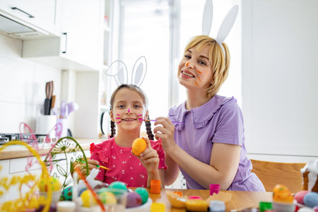 A mother and daughter wearing bunny ears are happily painting Easter eggs and applying face paint to...
