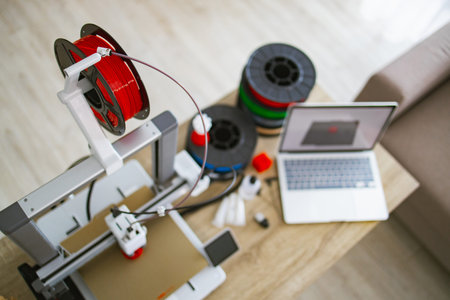 A 3D printer is set up on a wooden table with spools of filament and a laptop nearby, ready for...