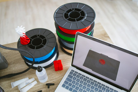 A laptop displays a 3D model next to spools of filament, glue, and tools, ready for additive...