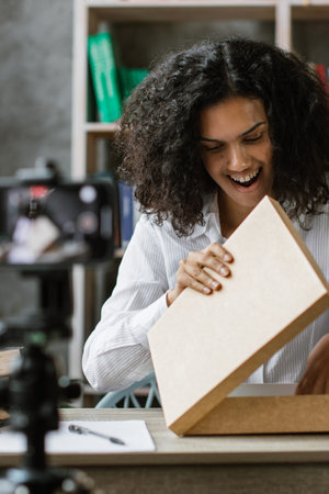 A young woman with curly hair excitedly opens a package while recording a video on her phone, capturing the moment of discovery