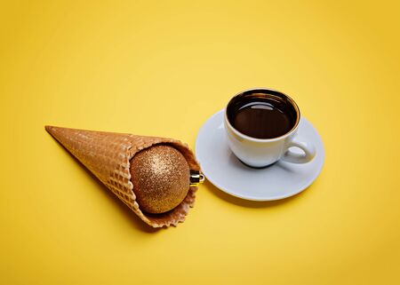 Espresso coffee mug and waffle cup with golden sparkling christmas ball in ice cream waffle cone on yellow background.の写真素材