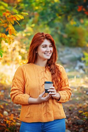 Red-haired smiling woman with a glass of coffee in her hands in the autumn park.の写真素材