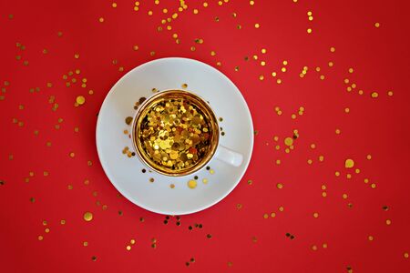Gold sparkles in a small cup with a gold border on a white saucer on the red background.の写真素材