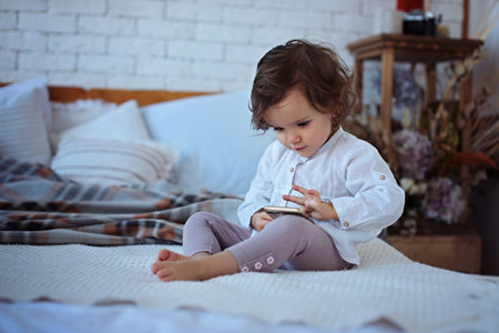Little girl sitting on the bed holding a smartphone, playing games and watching cartoons. Baby...