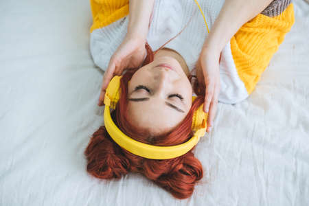 A female with red hair in yellow headphones lies on the bed and listens to music. View from above.の写真素材