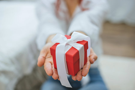 Red gift box with white ribbon in female hands. Close-up. A girl in a white shirt with a gift box in...