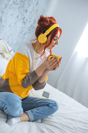 A girl with red bright hair sits on the bed, drinks coffee and listens to music in yellow headphones.の写真素材