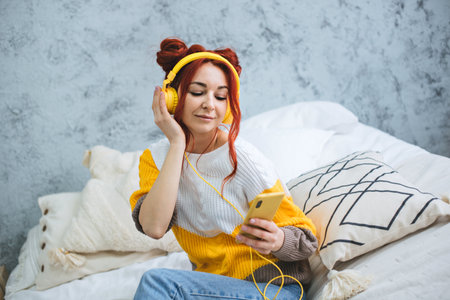 A girl with red bright hair sits on the bed and listens to music in yellow headphones.の写真素材